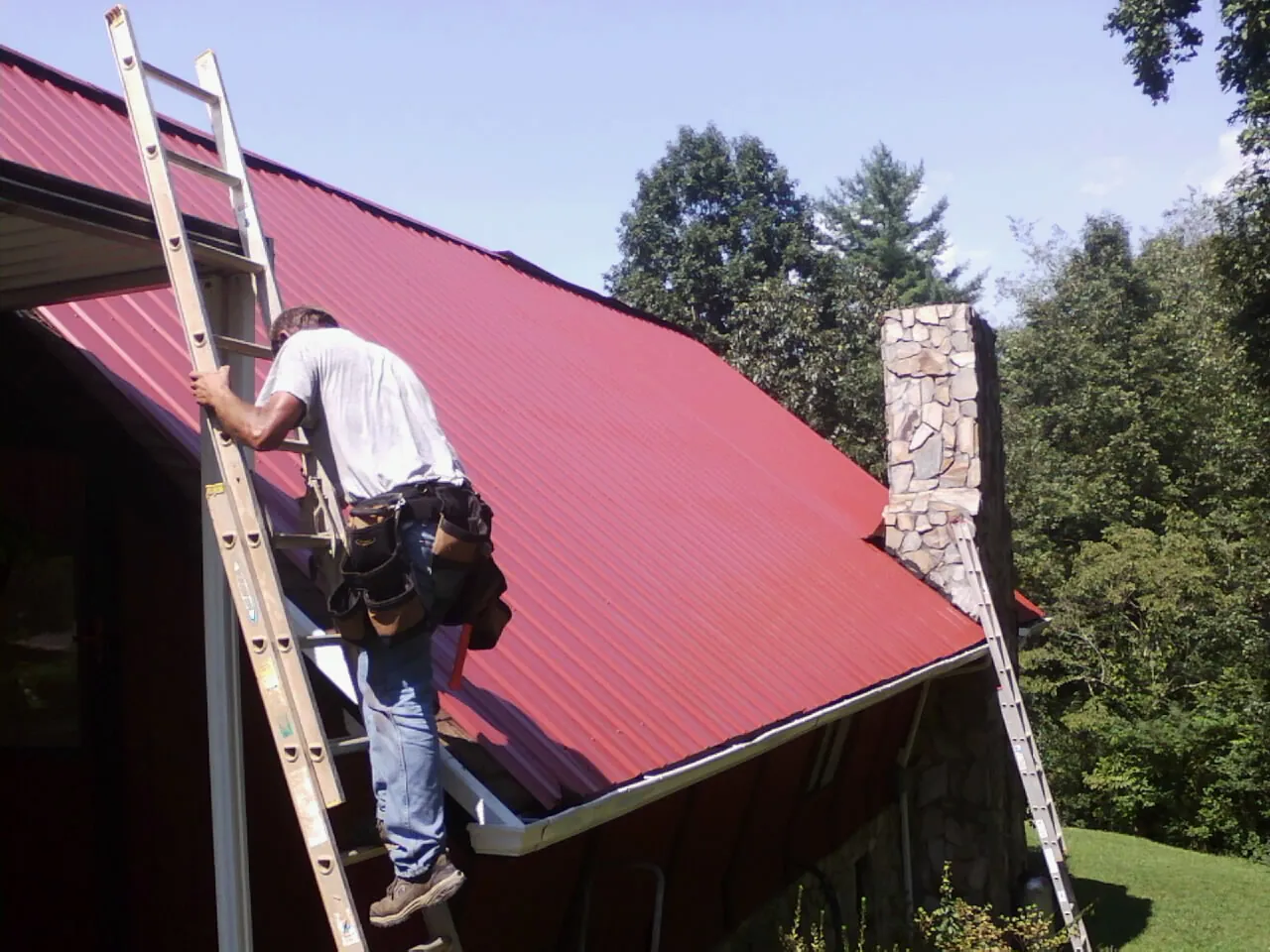 Roofer on a ladder beside a red metal roof and stone chimney — Soffit & Fascia Repair by Roofing Brothers in Barwick
